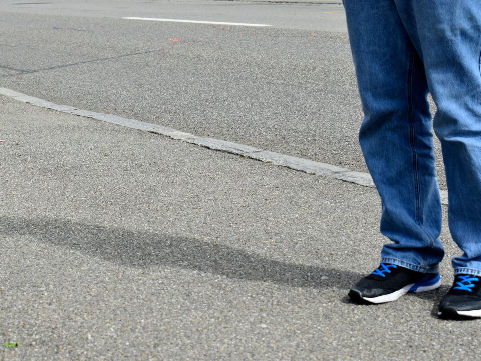 Ein Mann steht auf der Strasse. Es sind nur seine Füsse, Beine und Schatten seines Körpers auf dem Boden zu sehen.